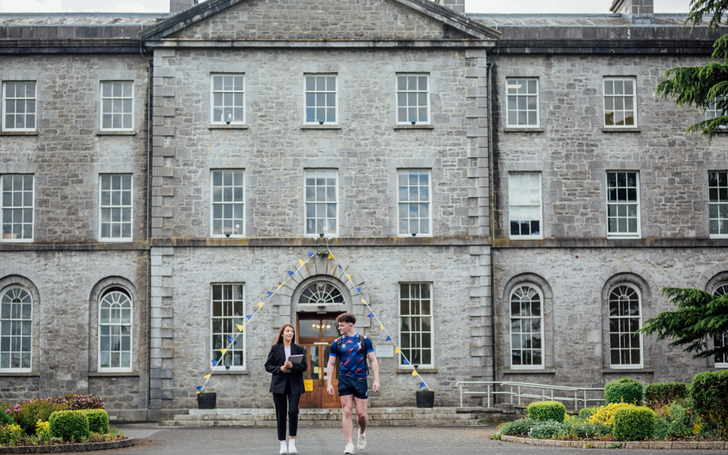 A female and female student are pictured walking together. There is a large grey 19th century building in the background.