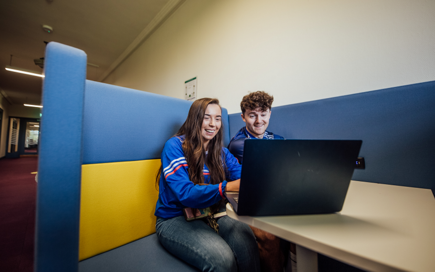 A female and male student are pictured sitting at a desk looking into a laptop screen.