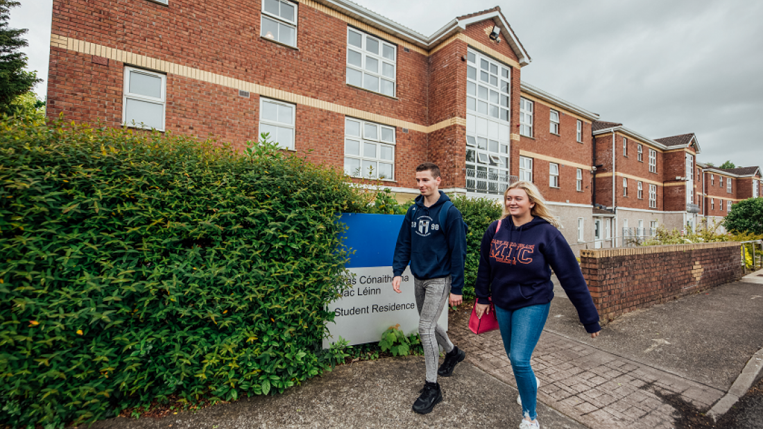 A male and female student walking on a footpath outside a large red brick building and green hedge.