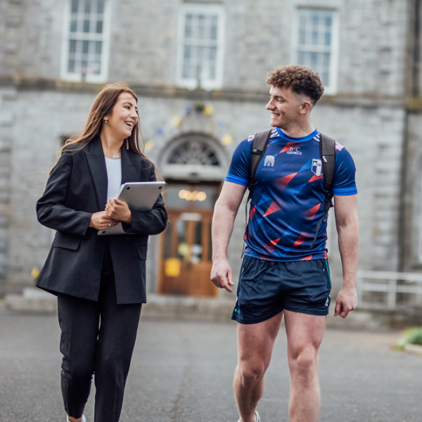 A female and male student are pictured walking side by side and chatting to each other. There is a grey 19th century building in the background with large windows.