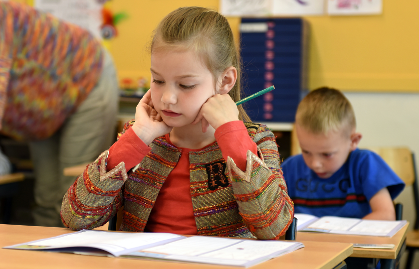 Girl pictured sitting at a table in a classroom with a boy sitting at another desk behind her