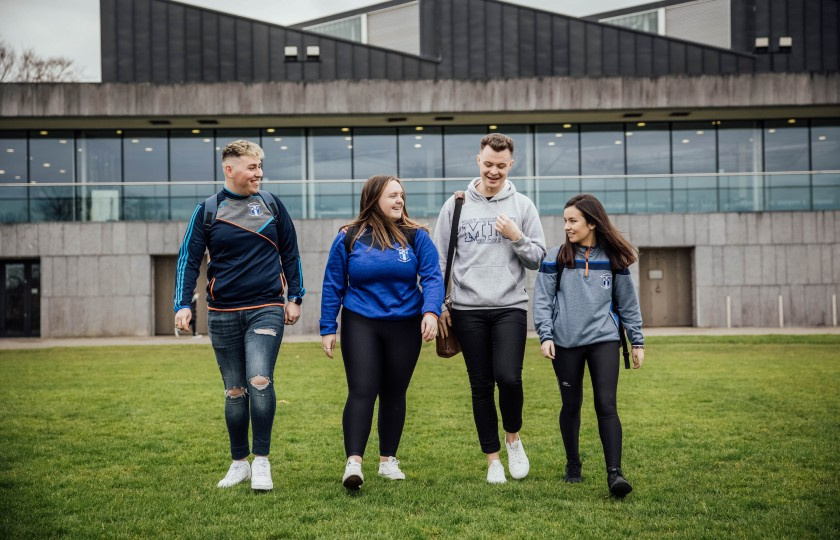 Four students walking across the playing field in front of MIC sports centre.