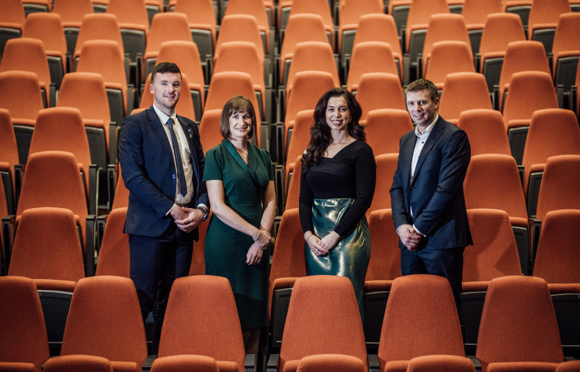 Declan, Petula, Miriam and Tomás standing in a theatre with orange chairs