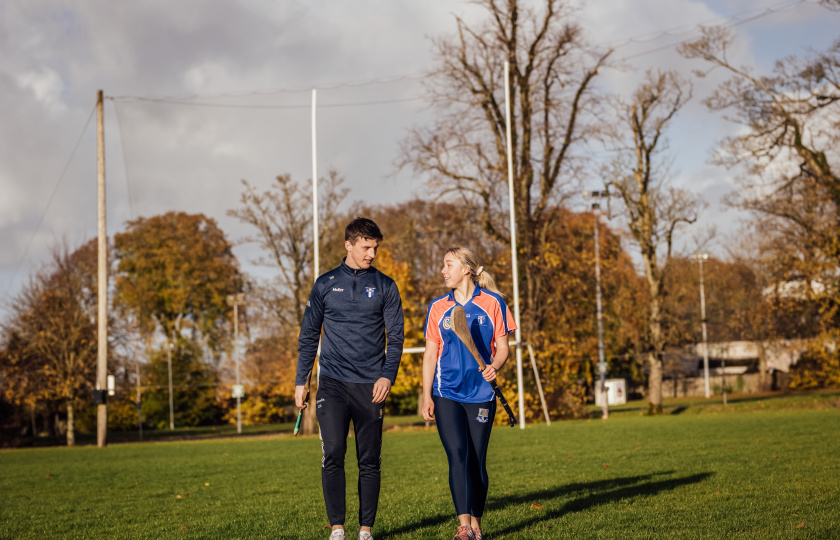 Diarmuid Ryan and Niamh Murphy walking on a GAA pitch and holding hurleys