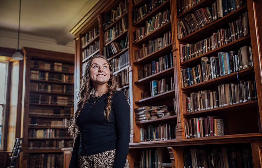 A female student standing in front of a large bookcase at the library.